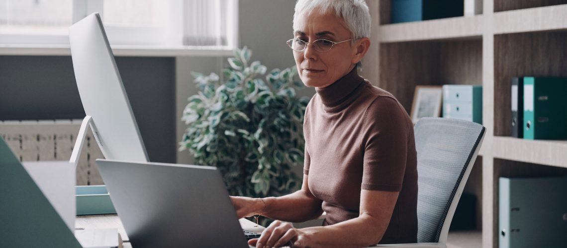 Confident senior businesswoman working at two computers while sitting in the office